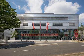 This picture shows building four of the new campus Derendorf of Hochschule Düsseldorf. The front of the building is of glass and steel, indoors you see red elements like the stairs and the walls. This building includes the lecture rooms.